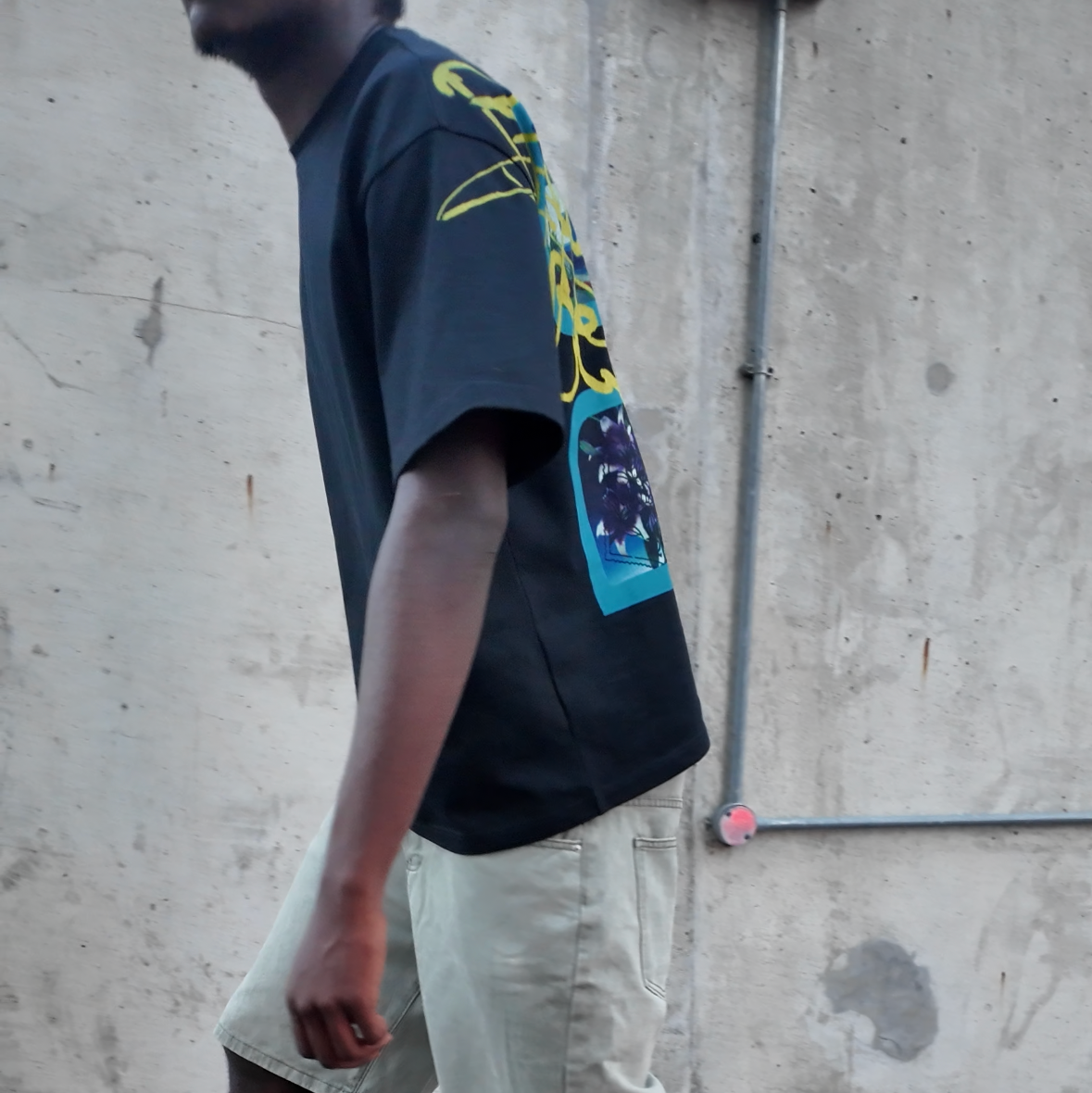 model wearing a black t-shirt, light shorts, and black sneakers on a concrete surface.